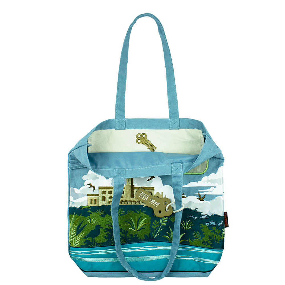 Blue tote bag with an embroidered design of Alcatraz Island, including prison buildings, plants, birds, and fog.
