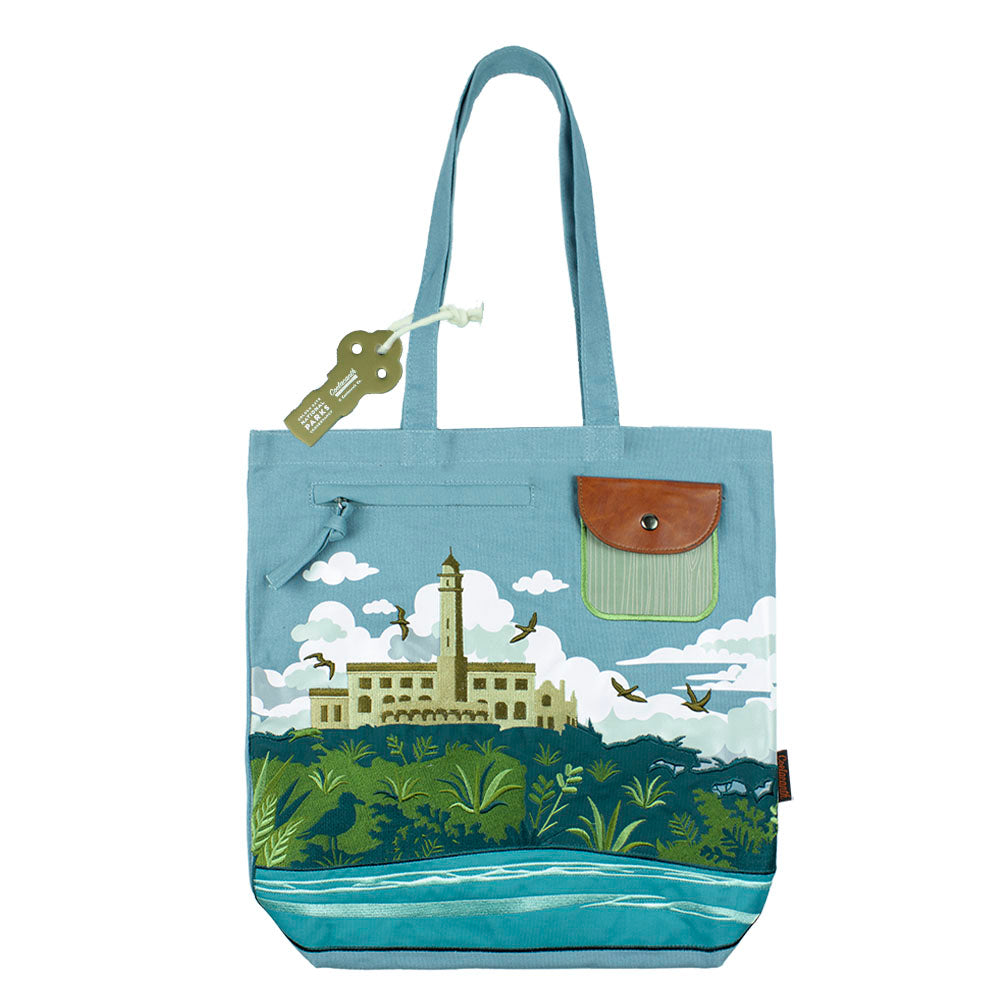 Blue tote bag with an embroidered design of Alcatraz Island, including prison buildings, plants, birds, and fog.