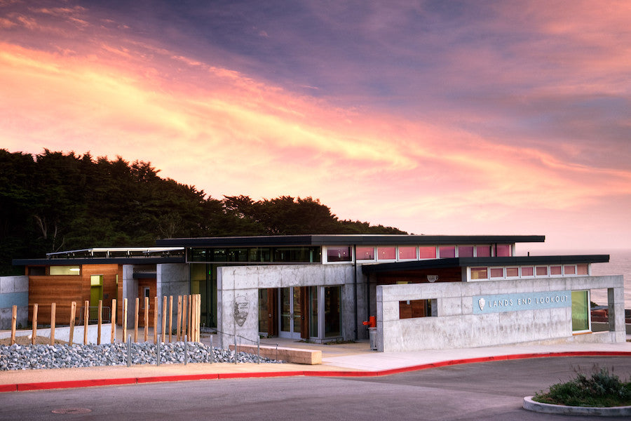 A dramatic view of the Lands End Lookout Visitor Center at sunset, with brightly lit pink, purple, and yellow clouds in the sky.